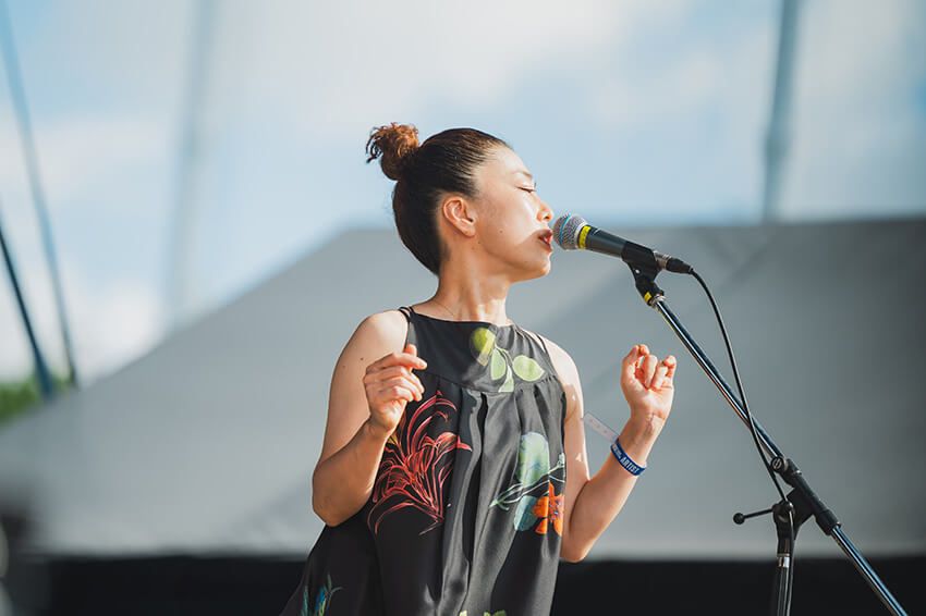 黒地に大ぶりの花柄があしらわれたワンピースに身を包み、マイクに向かって両手を広げる姿は圧倒的な存在感を放つ。力強くも柔らかい歌声は、ギターやベース、ドラム、そしてキーボードの多彩なサウンドと溶け合い、フェスならではの開放感をさらに加速させていった。 ©︎RHYTHM AND RIDE