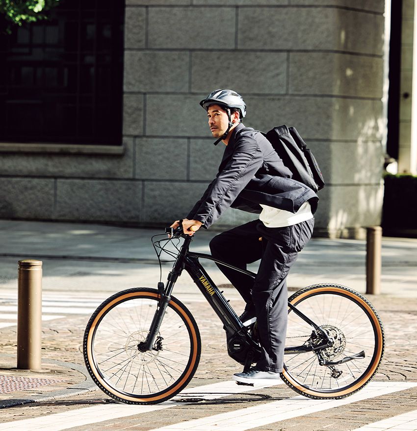 E-bike通勤なら、オフィスへ向かう満員電車のストレスから解放され、ペダルを漕いで心身ともに健やかな一日が始まる。オフロード対応のサスペンションフォークが、通勤途中の段差や凹凸のある道から伝わる衝撃を和らげ、街中でもその優れた性能を実感できる。さらにやや太めのブロックタイヤが、雨上がりの濡れた路面でも確かなグリップ力を発揮する。ジャケット4万7300円、パンツ3万800円／ともにナナミカ（ナナミカ 代官山　03-5728-6550）、バッグ9万6800円／ブリーフィング（ブリーフィング 六本木ヒルズ店　03-6434-5922）、スニーカー1万9250円／ソレル（コロンビアスポーツウェアジャパン　0120-193-803）